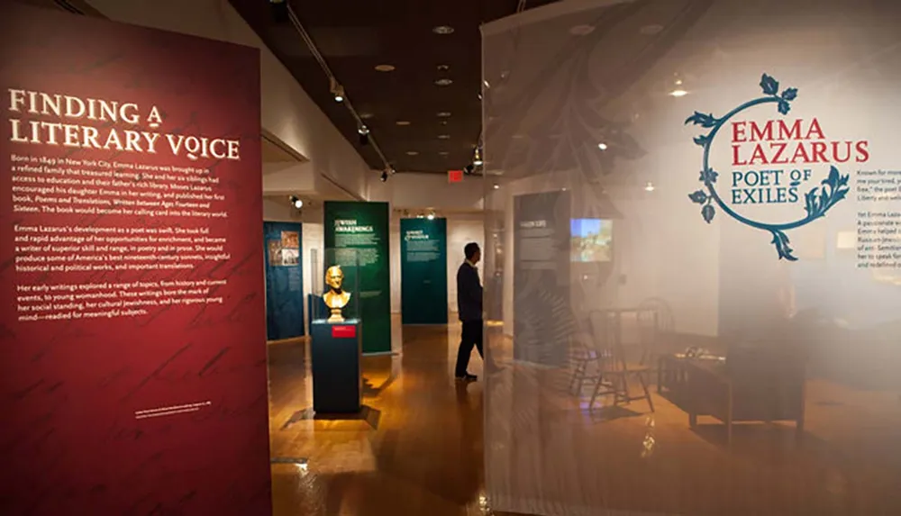 The image shows an interior view of an exhibition space dedicated to Emma Lazarus featuring informational panels and a bust of the poet with an individual observing the display