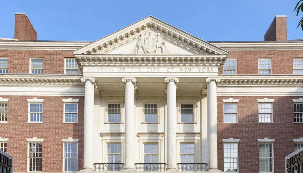The image shows the grand facade of the Museum of the City of New York characterized by its stately columns and classic brickwork under a clear sky