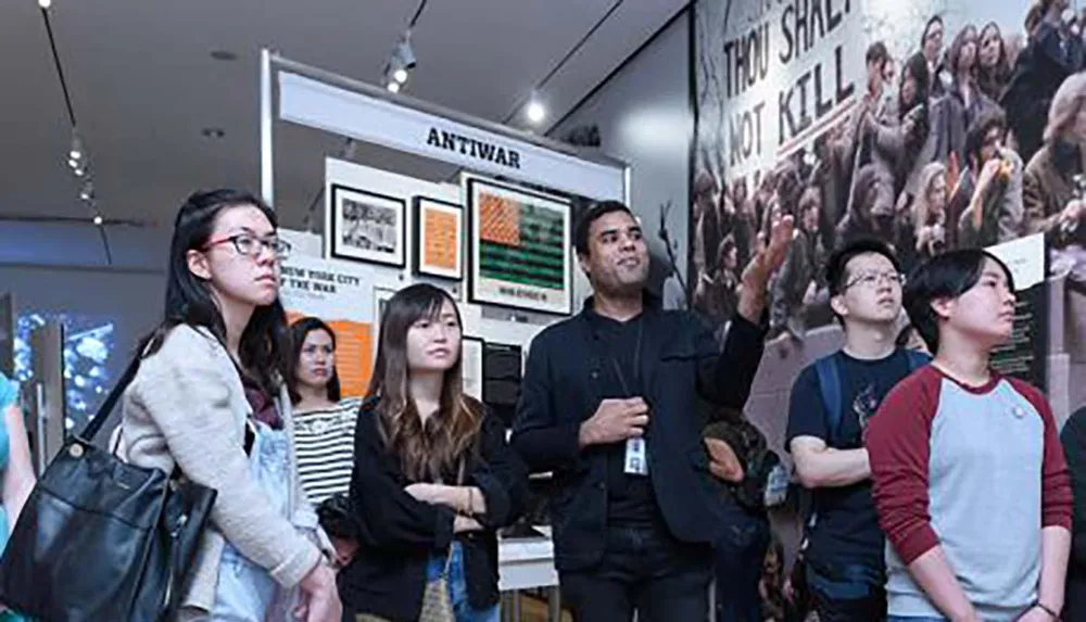 A group of people are attentively observing and listening to a presentation in a gallery with posters and images on the walls including one with the words THOU SHALT NOT KILL