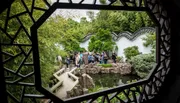The image shows a group of people exploring a traditional Chinese garden, framed through an ornate, geometrically patterned window.