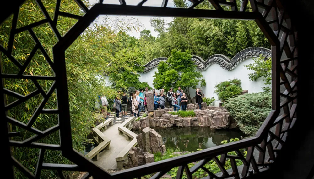 The image shows a group of people exploring a traditional Chinese garden framed through an ornate geometrically patterned window