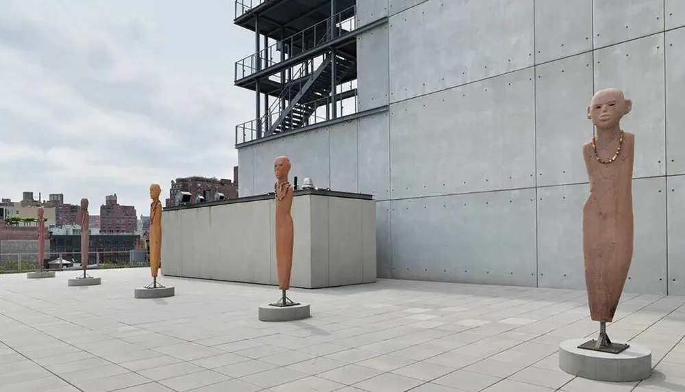 The image shows a series of elongated abstract humanoid sculptures displayed on an open terrace against an overcast sky and industrial backdrop