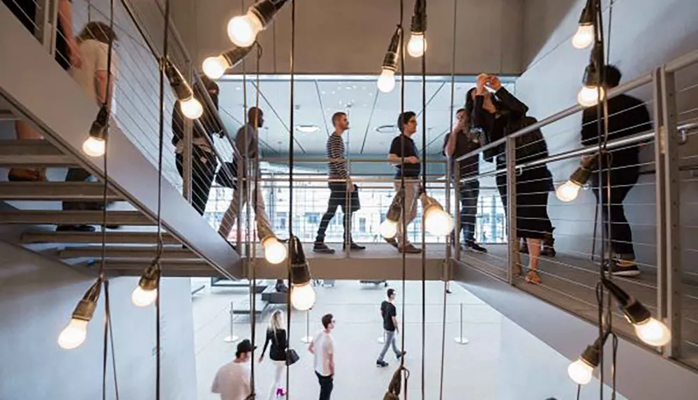 People are interacting on various levels of a modern open staircase adorned with hanging light bulbs in a contemporary interior space