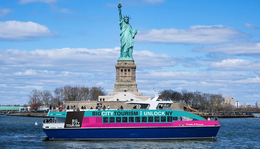 A colorful tour boat is passing in front of the Statue of Liberty under a partly cloudy sky