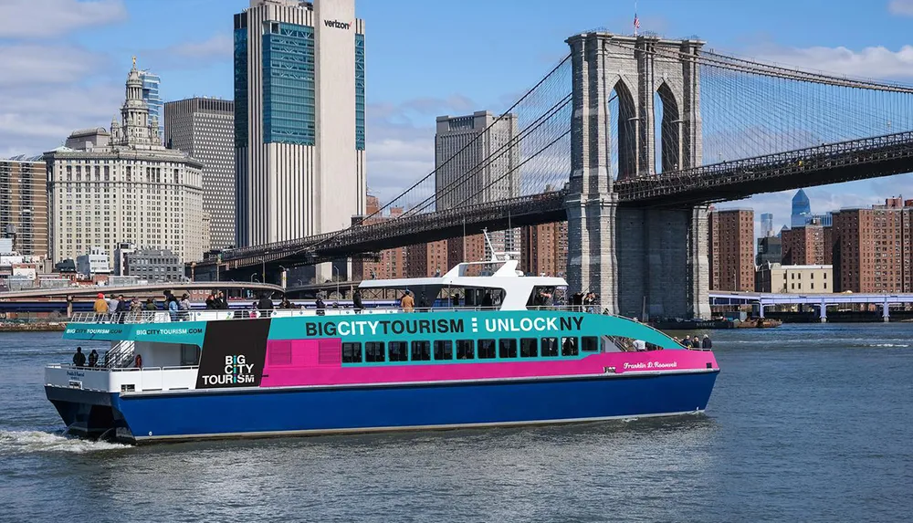 A colorful tourist boat sails on the water in front of the Brooklyn Bridge with a backdrop of Manhattan skyscrapers