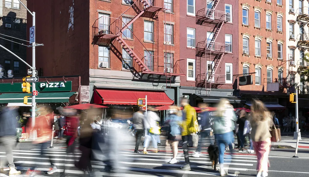 Pedestrians in motion cross a sunlit urban street corner featuring a pizza shop under classic brick buildings with fire escapes