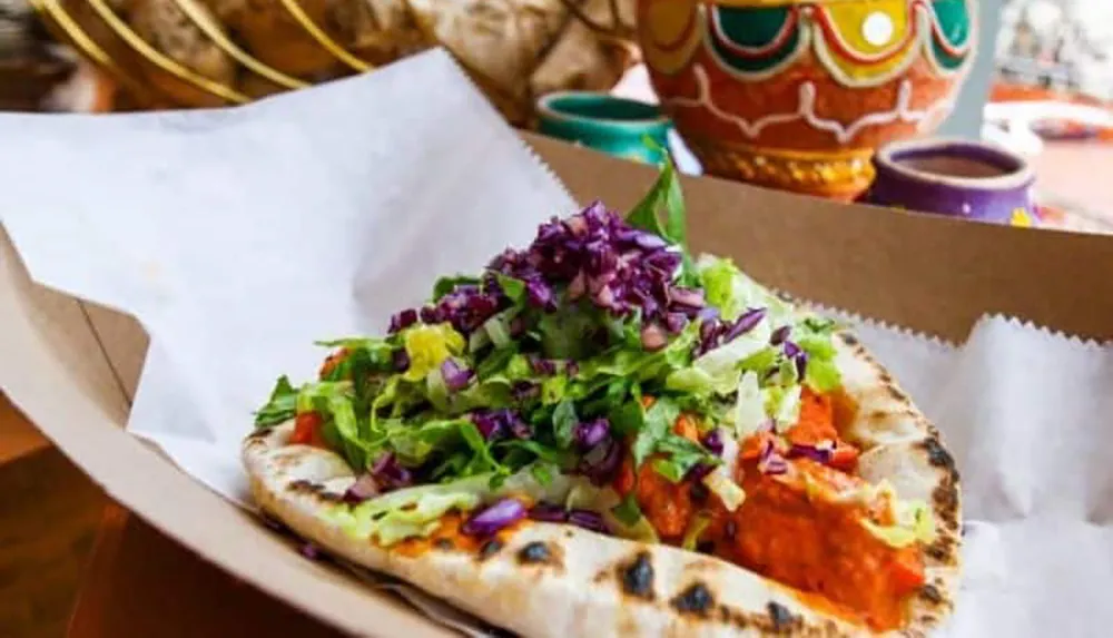 The image shows a vibrant and appetizing stuffed pita bread overflowing with fresh salad and what appears to be a red possibly spicy protein served in a casual dining environment