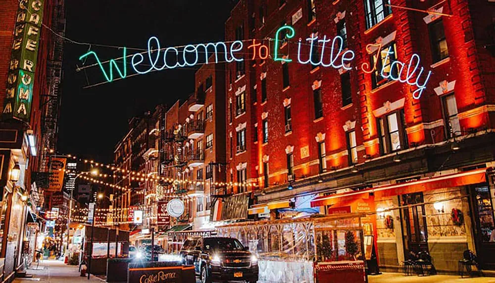 This image shows a neon sign saying Welcome to Little Italy above a lively street scene at night adorned with string lights and lined with restaurants and shops