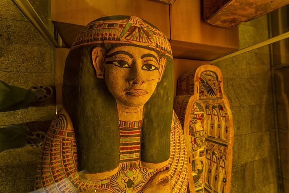 The image shows a colorful ornate Egyptian sarcophagus displayed in a shadowy room reflecting ancient funerary art