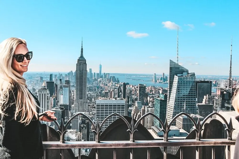 A woman in sunglasses is smiling at the camera from a high vantage point overlooking the New York City skyline with the Empire State Building prominently in the background