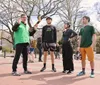 Four individuals are standing in a park-like setting with the person on the left appearing to speak and gesture to the others who are attentively listening