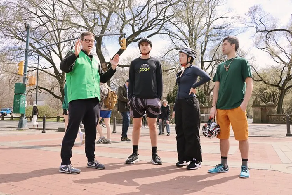 Four individuals are standing in a park-like setting with the person on the left appearing to speak and gesture to the others who are attentively listening