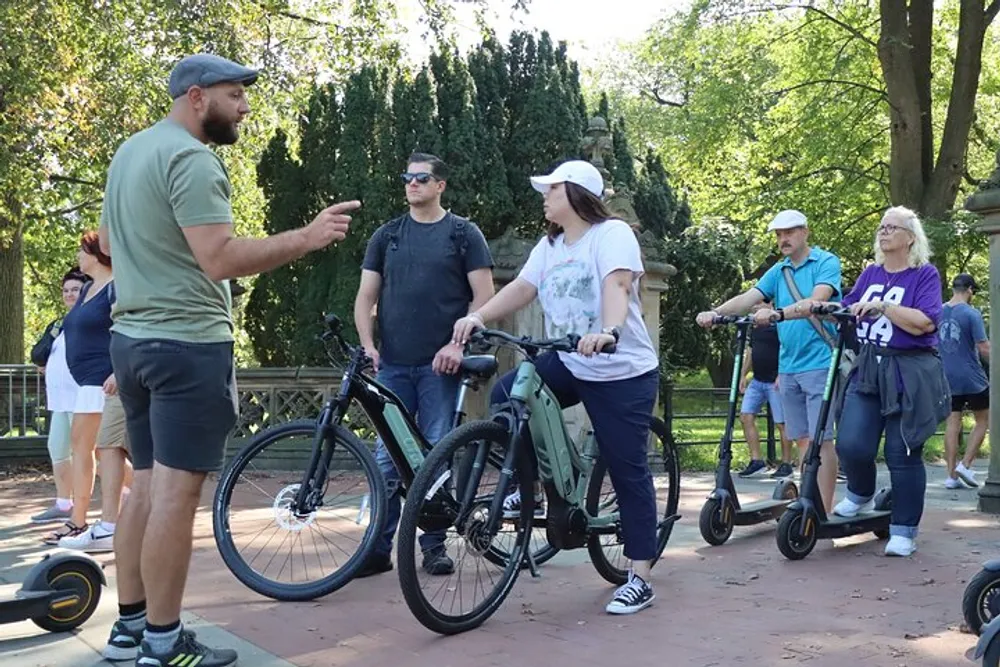 A group of people with bicycles and electric scooters appear to be having a conversation or receiving instructions outdoors on a sunny day