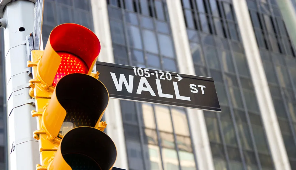 A red traffic light is illuminated above a street sign indicating Wall Street with directional arrows and building facades in the background