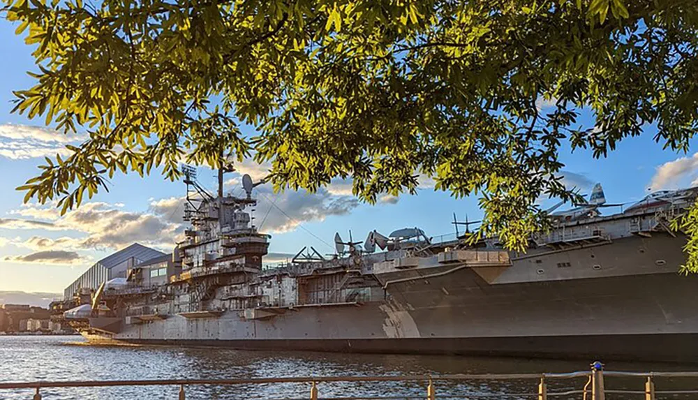 A large navy ship is docked by the waterfront with the setting sun casting warm light on its hull as seen through the branches of a tree