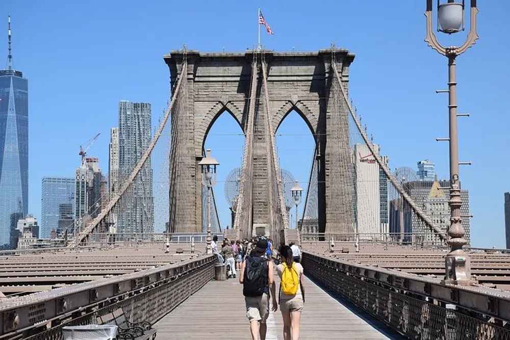 Pedestrians walk across the Brooklyn Bridge with its distinctive Gothic arches against the backdrop of the New York City skyline on a clear day