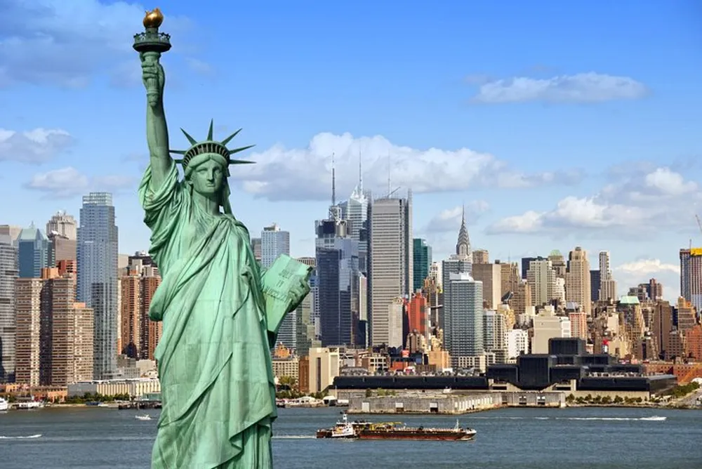 The Statue of Liberty stands in the foreground with the skyline of New York City in the background under a partly cloudy sky