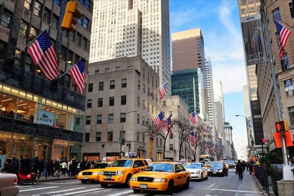 Iconic yellow taxis navigate a bustling downtown street lined with American flags and skyscrapers under a clear blue sky