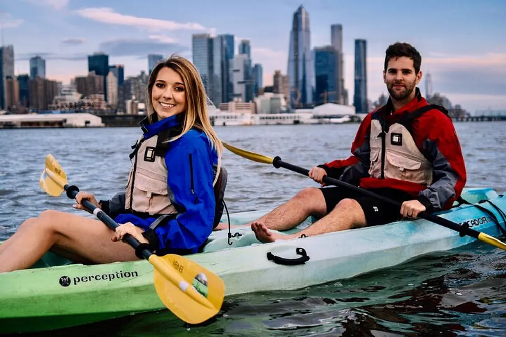 Two people are smiling and kayaking together with a city skyline in the background at dusk