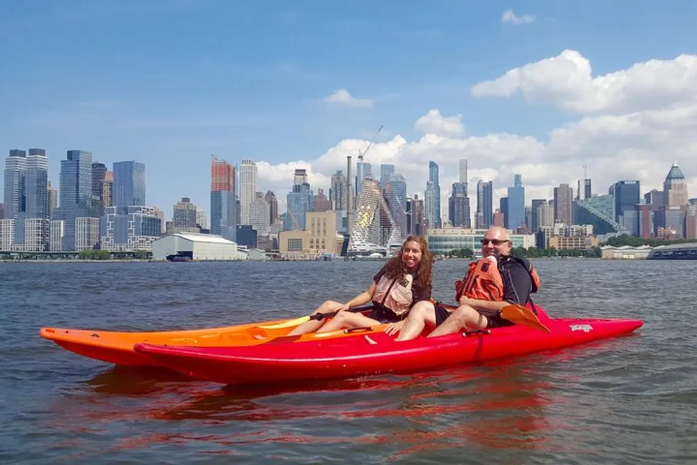 Two people are kayaking on a river with a city skyline in the background under a partly cloudy sky