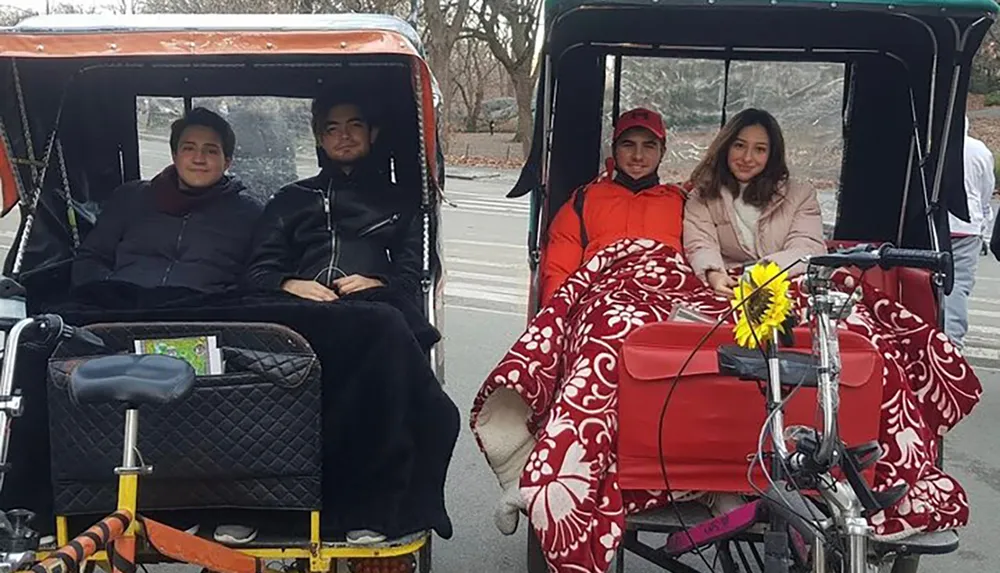 Four people are smiling while sitting in parked pedicabs with blankets with a sunflower affixed to one of the pedicabs suggesting they are on a leisurely outing