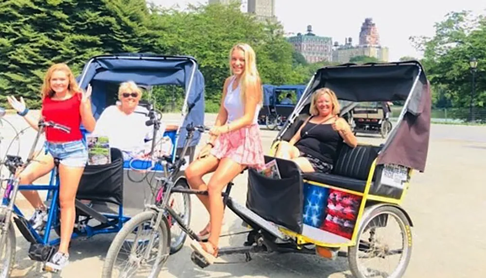 Two pedicabs each with a passenger are parked side by side with a woman standing by one and a driver sitting on the other all smiling for a photo in a sunny park-like setting