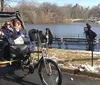 Three passengers are enjoying a ride in a pedal-powered rickshaw while a photographer captures the scene in a park with snow patches on the ground and a lake in the background