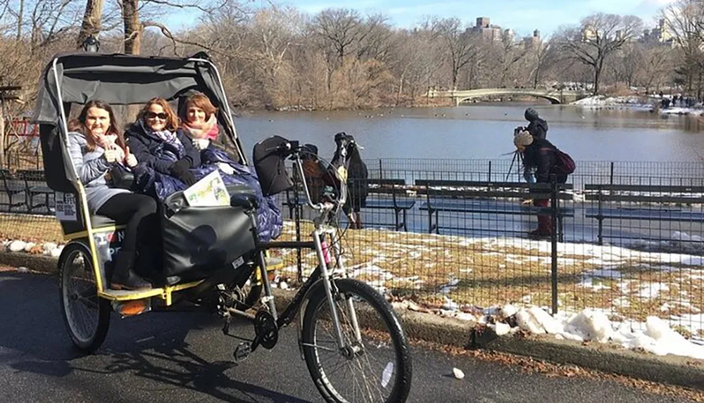 Three passengers are enjoying a ride in a pedal-powered rickshaw while a photographer captures the scene in a park with snow patches on the ground and a lake in the background