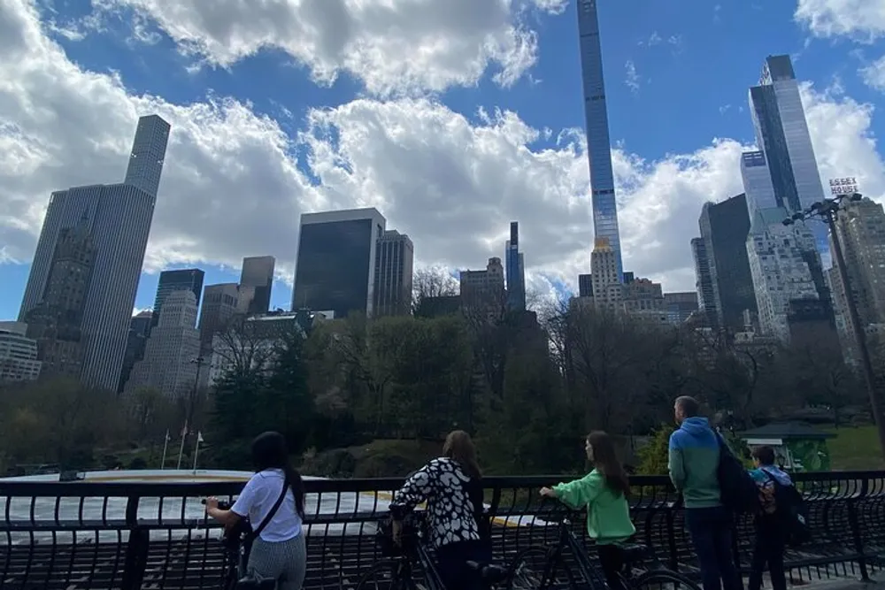 People are enjoying a view of a city skyline from a park with towering skyscrapers under a partly cloudy sky