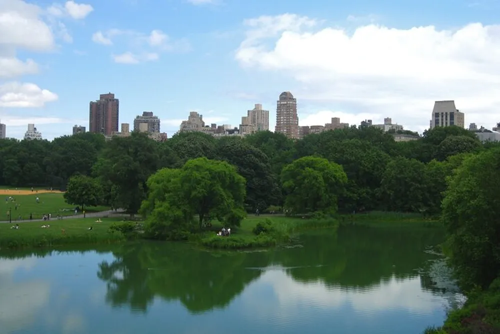 A lush green park with a tranquil pond in the foreground and a city skyline in the background under a partly cloudy sky