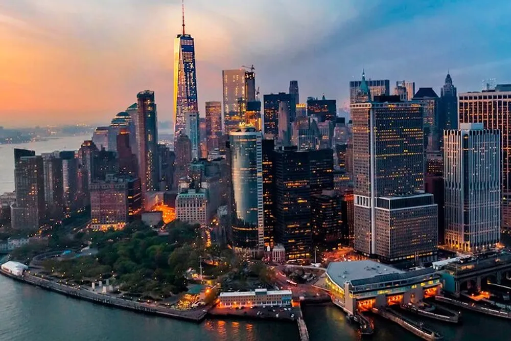 An aerial view captures the illuminated skyline of Lower Manhattan at dusk with the Hudson River in the background