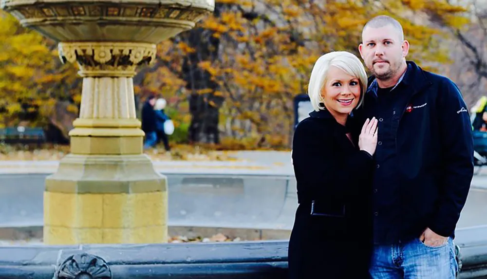 A smiling woman in a black coat poses with her arm around a man in front of an ornate fountain in a park with autumn foliage