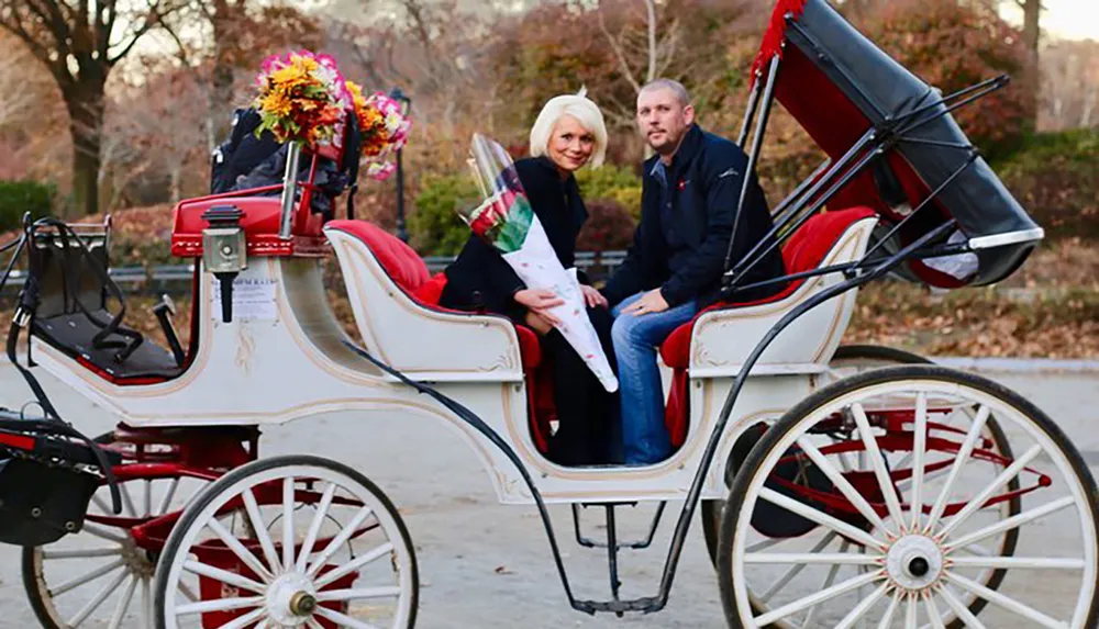 A couple sits in a white horse-drawn carriage with the woman holding a bouquet of flowers suggesting a romantic or celebratory outing