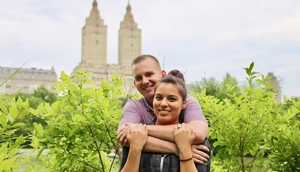 A smiling couple poses for a photo with one person standing behind the other wrapping their arms around them with a backdrop of lush greenery and tall historic-looking buildings