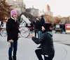 A person is kneeling on one knee in what appears to be a marriage proposal to a pleasantly surprised individual in a park with onlookers and horse-drawn carriages in the background