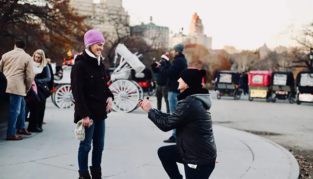 A person is kneeling on one knee in what appears to be a marriage proposal to a pleasantly surprised individual in a park with onlookers and horse-drawn carriages in the background