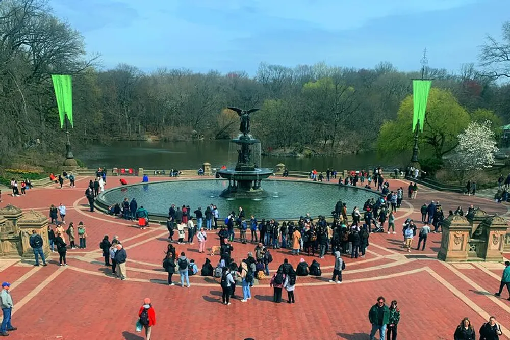 This image shows a bustling scene of people gathered around the Bethesda Fountain in Central Park New York City with the terrace overlooking the water on a clear day