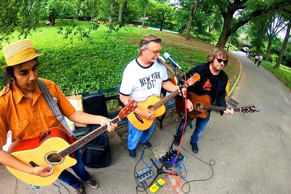 Three musicians are performing outdoors with acoustic guitars and a microphone surrounded by greenery in what appears to be a park setting