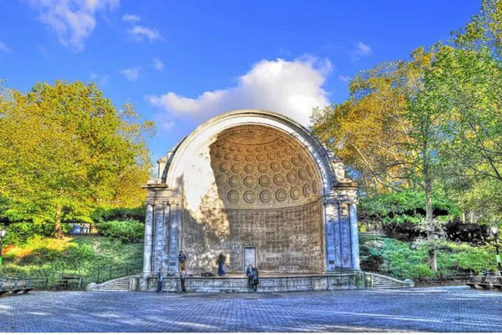 The image shows the Naumburg Bandshell located in Central Park New York with people enjoying a sunny day amidst the surrounding vibrant greenery