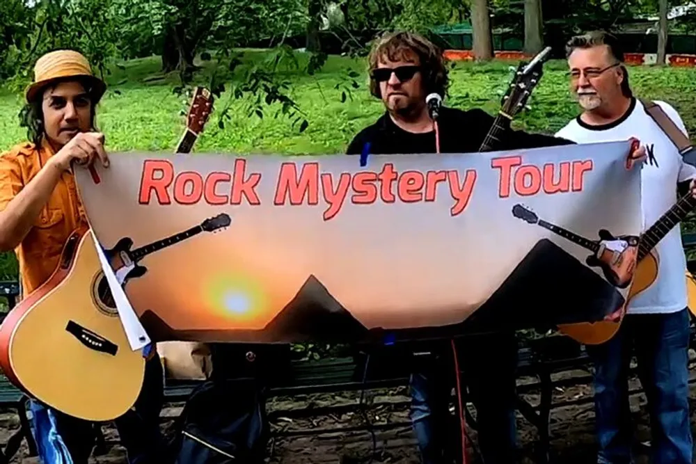 Three musicians are holding a banner that reads Rock Mystery Tour with two of them holding guitars