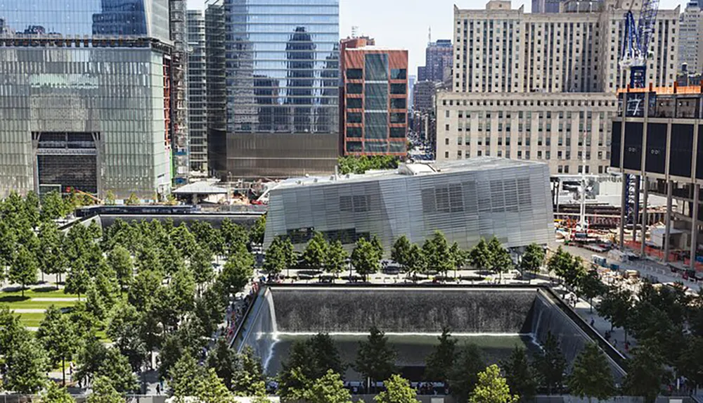 The image shows a modern urban park with rows of trees surrounding a reflective memorial pool bordered by contemporary buildings in a bustling cityscape