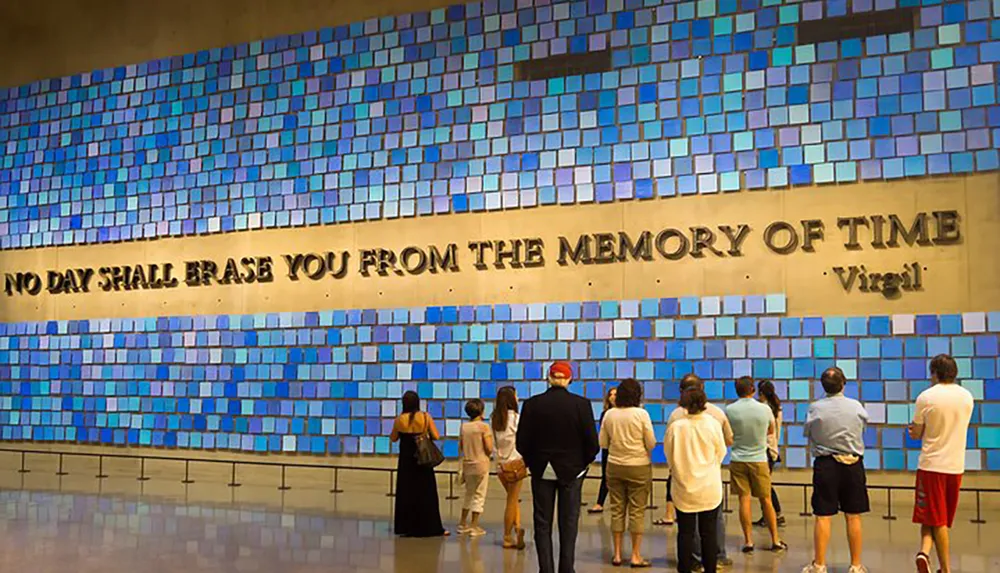 Visitors are standing in front of a large wall with blue squares and a quote that reads NO DAY SHALL ERASE YOU FROM THE MEMORY OF TIME - Virgil