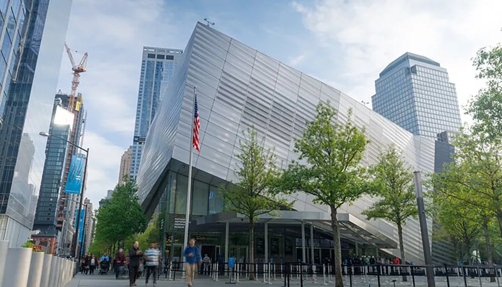 This image shows a modern building with an angled facade flanked by American flags amidst a cityscape with people walking along the sidewalk