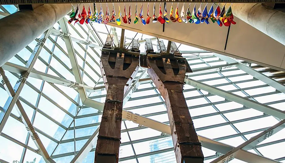 The image shows two preserved columns from a damaged structure displayed in a modern space with a glass ceiling beneath which hangs a row of colorful firefighter helmets