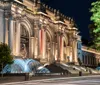 The image shows a grand illuminated neoclassical building at night possibly a museum with a lit fountain in the foreground and people sitting on its steps