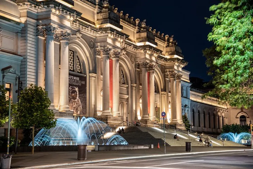 The image shows a grand illuminated neoclassical building at night possibly a museum with a lit fountain in the foreground and people sitting on its steps