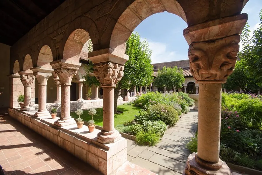 The image shows a serene cloister garden viewed from under an arched stone colonnade displaying lush greenery and medieval architectural details