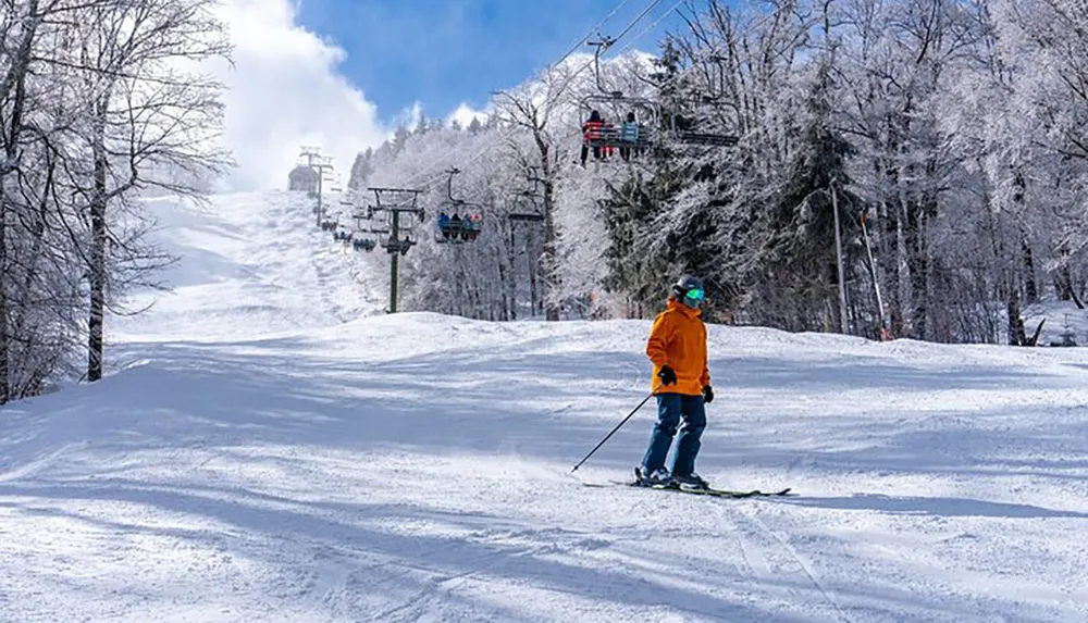 A skier in an orange jacket descends a snowy slope with a ski lift running overhead amidst a winter landscape