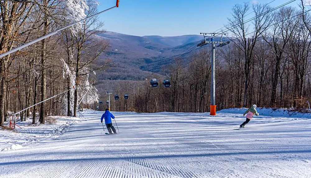 Skiers descend a groomed slope with a ski lift and mountainous background on a clear day