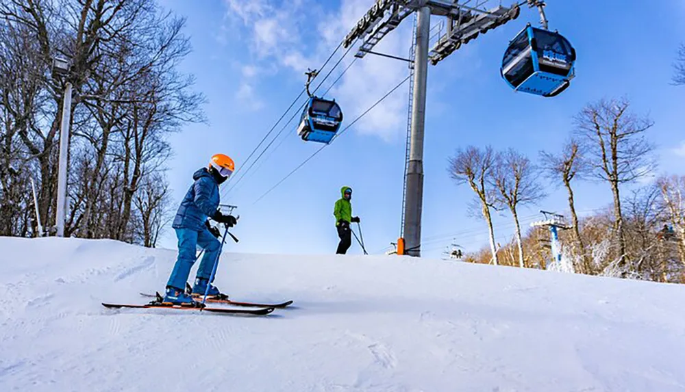 Two skiers are descending a snowy slope with a ski lift operating in the background under a clear blue sky